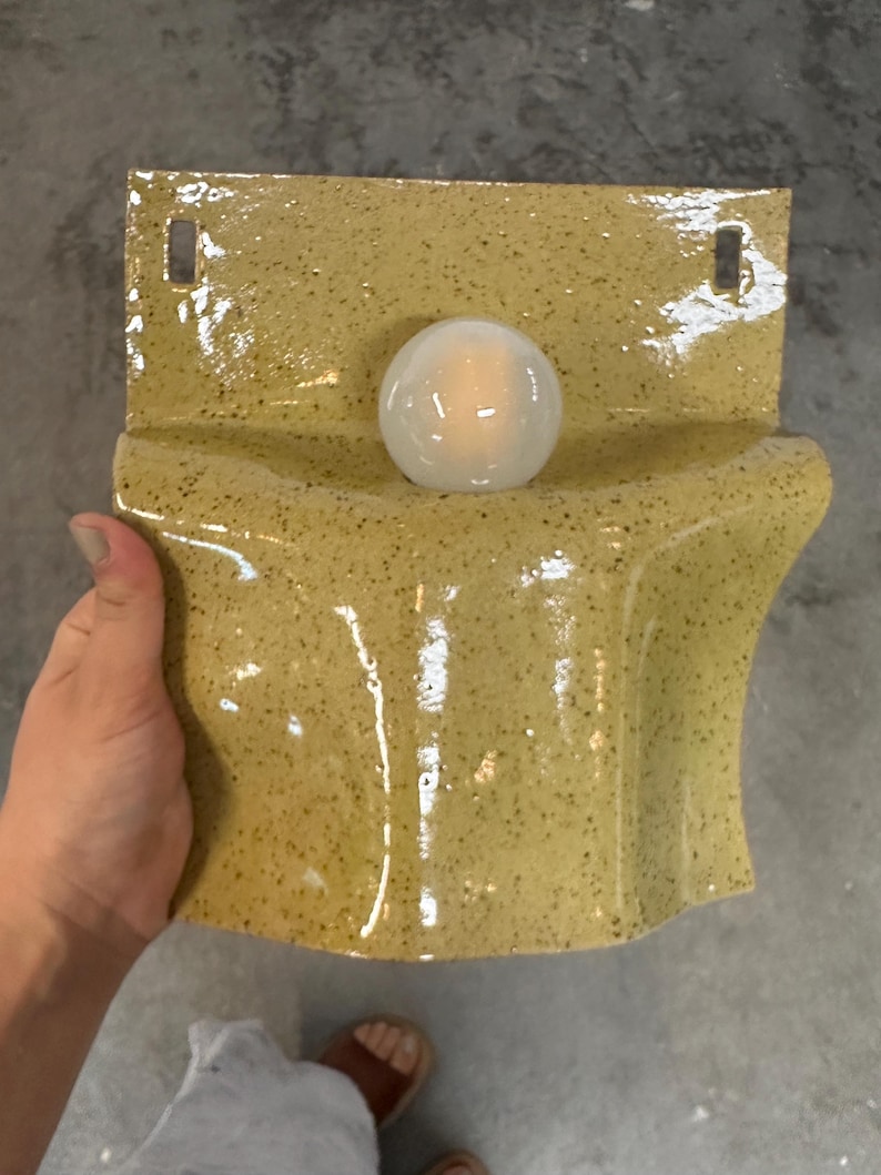 May include: A wall-mounted ceramic light fixture with a glossy, speckled mustard yellow glaze. The piece features a curved, wave-like form with two rectangular mounting holes at the top and a single, central white spherical light bulb.