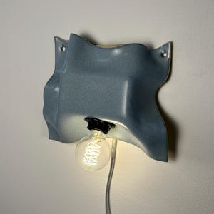 May include: A wall-mounted light fixture featuring a wavy, blue ceramic shade with a glossy finish. The fixture includes a black socket holding a clear, round Edison-style light bulb that emits a warm glow, connected by a thin, transparent power cord.