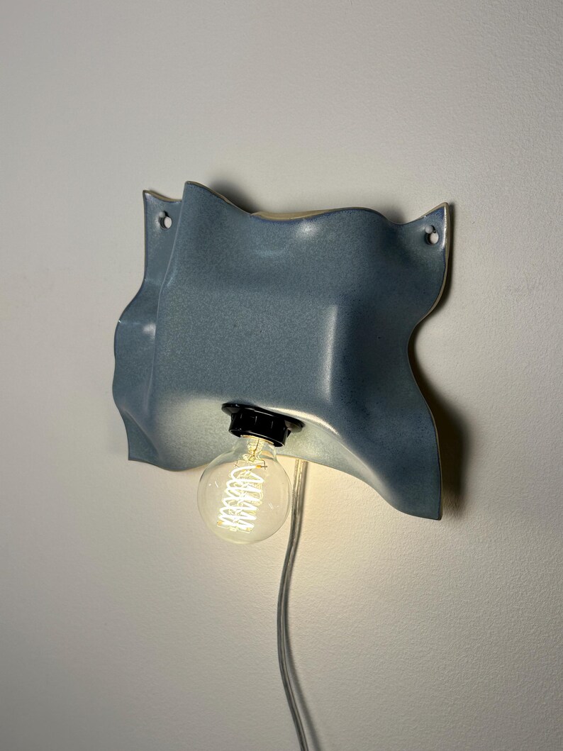 May include: A wall-mounted light fixture featuring a wavy, blue ceramic shade with a glossy finish. The fixture includes a black socket holding a clear, round Edison-style light bulb that emits a warm glow, connected by a thin, transparent power cord.