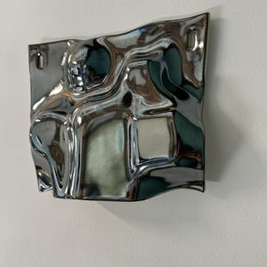 Pu&ograve; includere: Un oggetto decorativo da parete quadrato con una finitura cromata lucida altamente riflettente. La superficie presenta onde irregolari, fluide e ondulate che creano riflessi distorti. Include due piccole fessure di montaggio rettangolari.