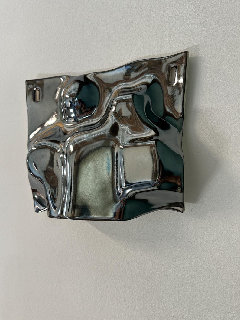 Pu&ograve; includere: Un oggetto decorativo da parete quadrato con una finitura cromata lucida altamente riflettente. La superficie presenta onde irregolari, fluide e ondulate che creano riflessi distorti. Include due piccole fessure di montaggio rettangolari.