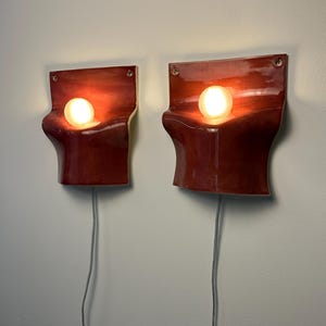May include: A pair of wall-mounted sconces featuring a glossy, deep red ceramic body with a curved, wave-like silhouette. Each fixture holds a single glowing spherical light bulb and includes a grey electrical cord extending from the bottom.