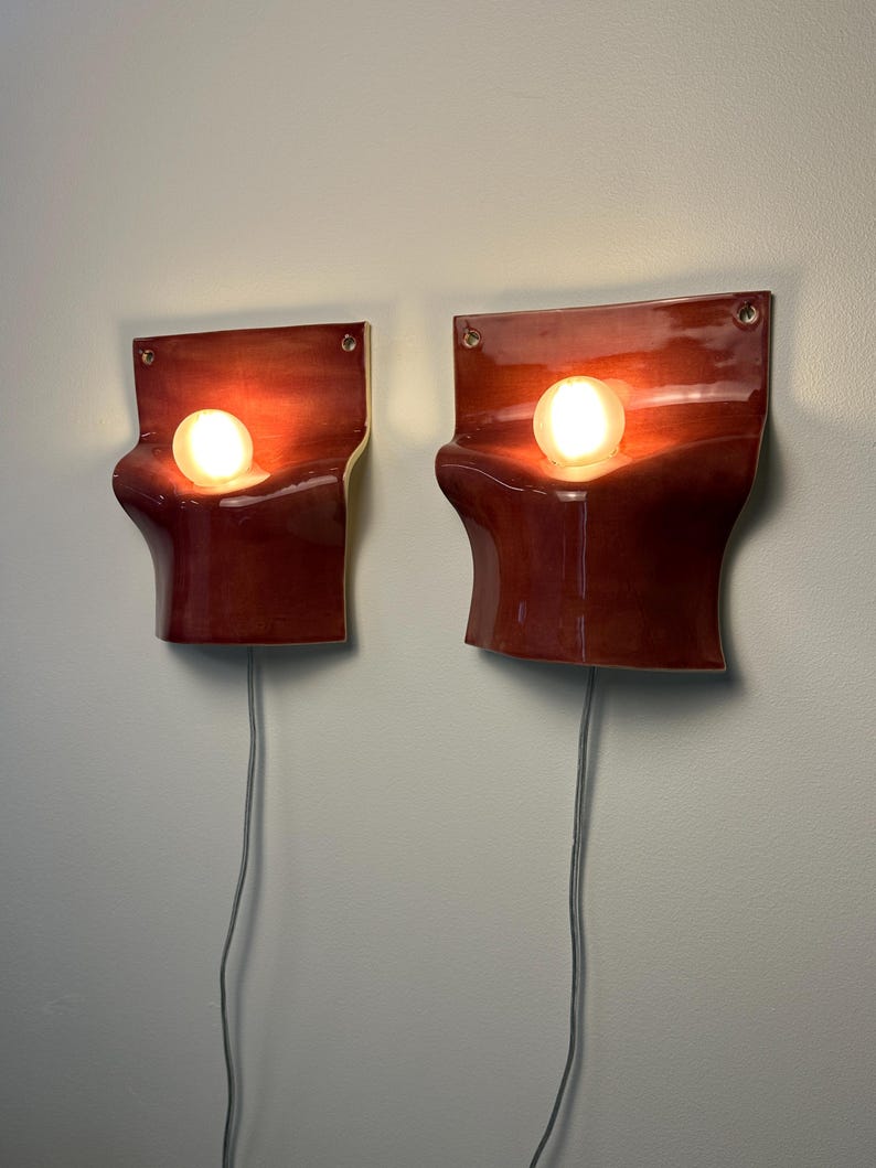 May include: A pair of wall-mounted sconces featuring a glossy, deep red ceramic body with a curved, wave-like silhouette. Each fixture holds a single glowing spherical light bulb and includes a grey electrical cord extending from the bottom.