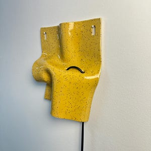 K&ouml;nnte beinhalten: Eine gelbe Wandleuchte aus Keramik mit schwarzem Sprenkelmuster. Das Objekt hat eine unregelm&auml;&szlig;ige, gefaltete und geschwungene Form mit zwei vertikalen Befestigungsschlitzen an der Oberseite und einem schwarzen Stromkabel an der Unterseite.