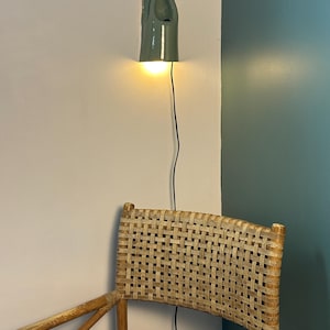 May include: A sage green ceramic wall sconce with a sculptural, elongated form and a downward-facing light source. The fixture features a smooth, glossy finish and is mounted on a wall, connected by a thin black electrical cord that hangs vertically.
