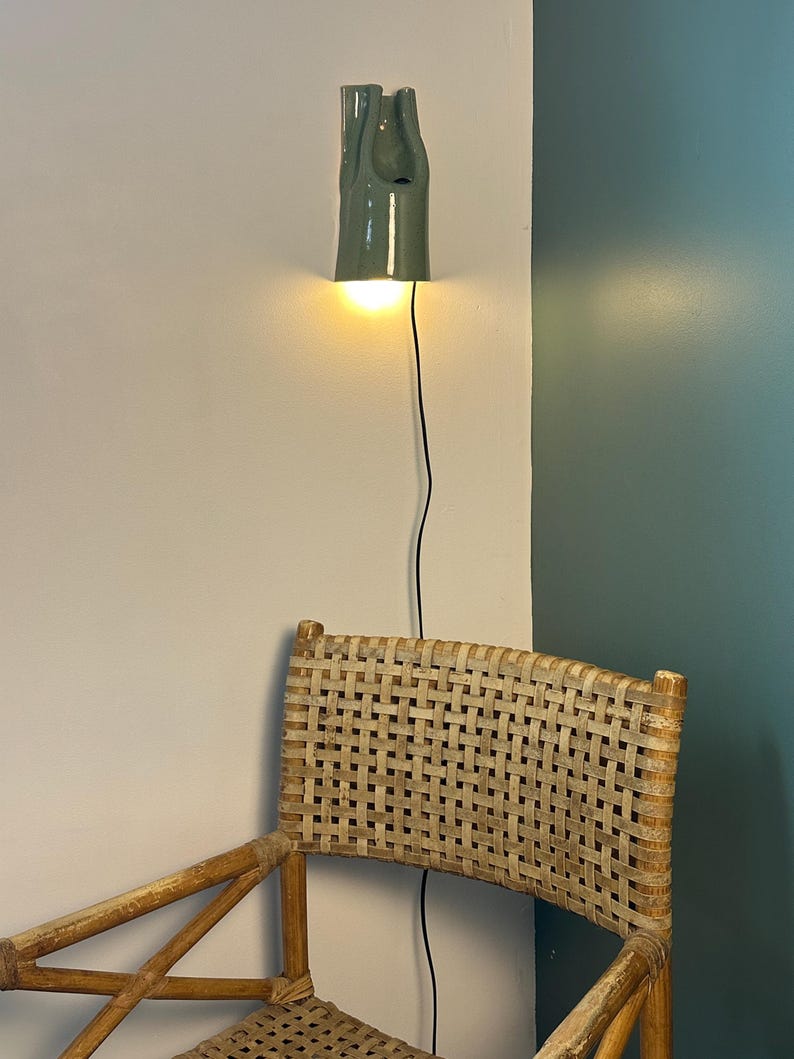 May include: A sage green ceramic wall sconce with a sculptural, elongated form and a downward-facing light source. The fixture features a smooth, glossy finish and is mounted on a wall, connected by a thin black electrical cord that hangs vertically.