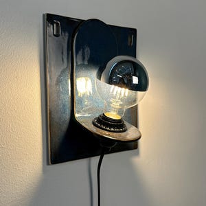 May include: A wall-mounted light fixture featuring a square, dark blue glazed ceramic backplate with a rounded arch design. A circular shelf holds a light bulb with a reflective silver top half, emitting a warm glow from the exposed filament base.