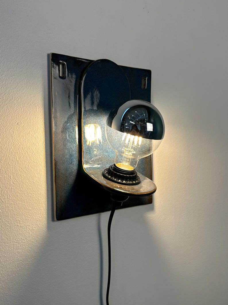 May include: A wall-mounted light fixture featuring a square, dark blue glazed ceramic backplate with a rounded arch design. A circular shelf holds a light bulb with a reflective silver top half, emitting a warm glow from the exposed filament base.