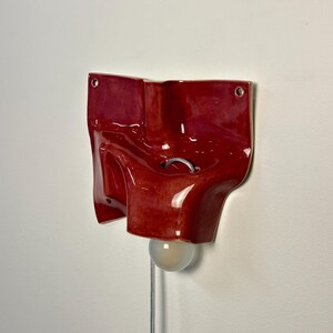 May include: A glossy, deep red ceramic wall sconce with an irregular, organic, folded shape. The fixture features two circular mounting holes at the top corners, a central metal loop, and a single round light bulb extending from the bottom.