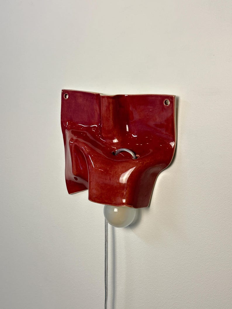 May include: A glossy, deep red ceramic wall sconce with an irregular, organic, folded shape. The fixture features two circular mounting holes at the top corners, a central metal loop, and a single round light bulb extending from the bottom.