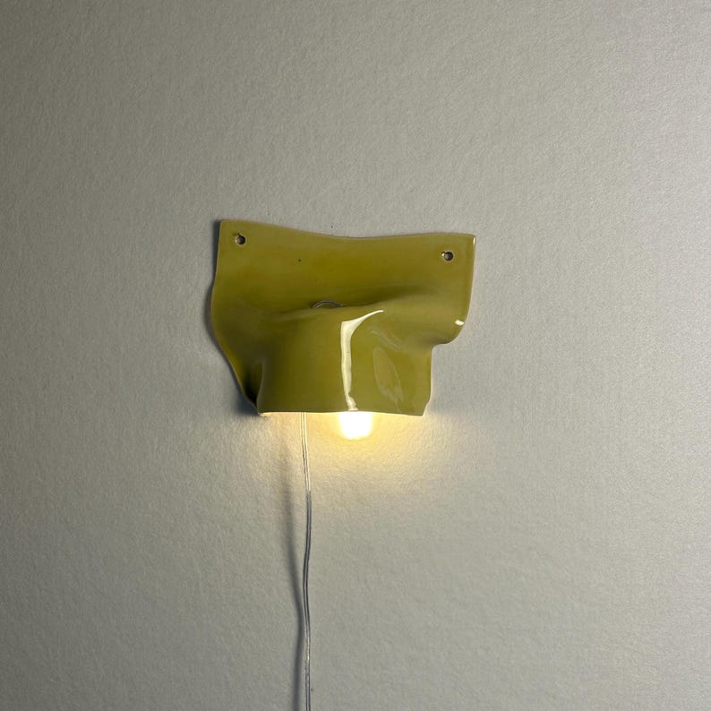 May include: A unique, wall-mounted light fixture. The fixture is a muted yellow-green color with a textured, organic shape. A small, round light bulb is visible at the bottom, emitting a warm glow. A thin, silver cord extends downward.