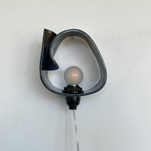 May include: A modern wall sconce with a unique, abstract design. The sconce features a dark blue, curved metal frame with a frosted bulb and black socket. A clear cord extends from the base, ready for plugging in.