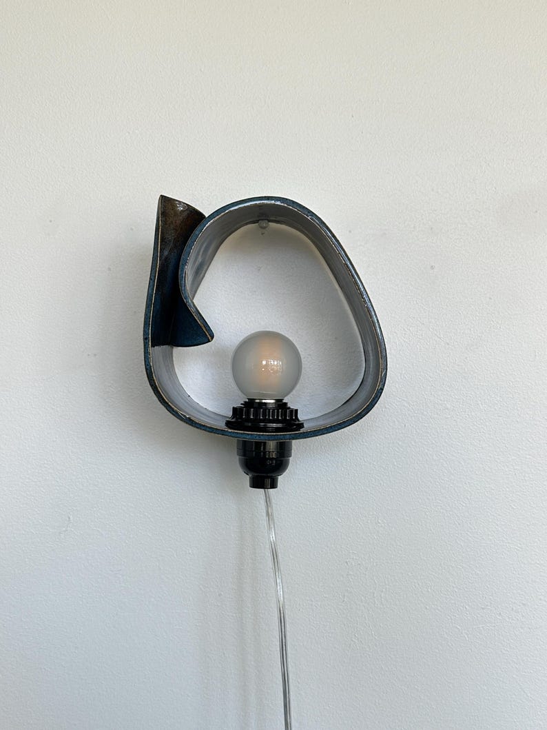May include: A modern wall sconce with a unique, abstract design. The sconce features a dark blue, curved metal frame with a frosted bulb and black socket. A clear cord extends from the base, ready for plugging in.
