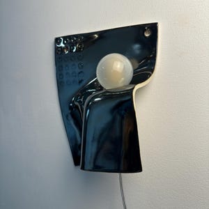 K&ouml;nnte beinhalten: Eine Wandleuchte mit einem gl&auml;nzend schwarzen Keramiksockel und einer strukturierten Oberfl&auml;che aus kleinen kreisf&ouml;rmigen Vertiefungen. Eine einzelne kugelf&ouml;rmige wei&szlig;e Glasgl&uuml;hbirne befindet sich in der Mitte der geschwungenen, unregelm&auml;&szlig;igen Keramikform.