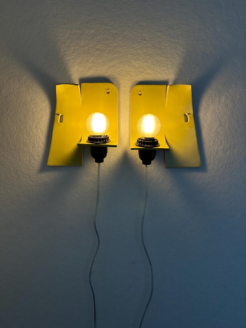 May include: A pair of wall-mounted sconces featuring bright yellow, flat, rectangular backplates with curved edges. Each sconce has a black socket holding a glowing, round, warm-toned light bulb, with a thin, clear power cord extending downward.