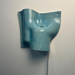 May include: A wall-mounted light blue ceramic wall light with a glossy finish and an organic, undulating shape. The fixture features two circular holes for mounting and a small silver-toned metal pull chain hanging from the centre for operation.