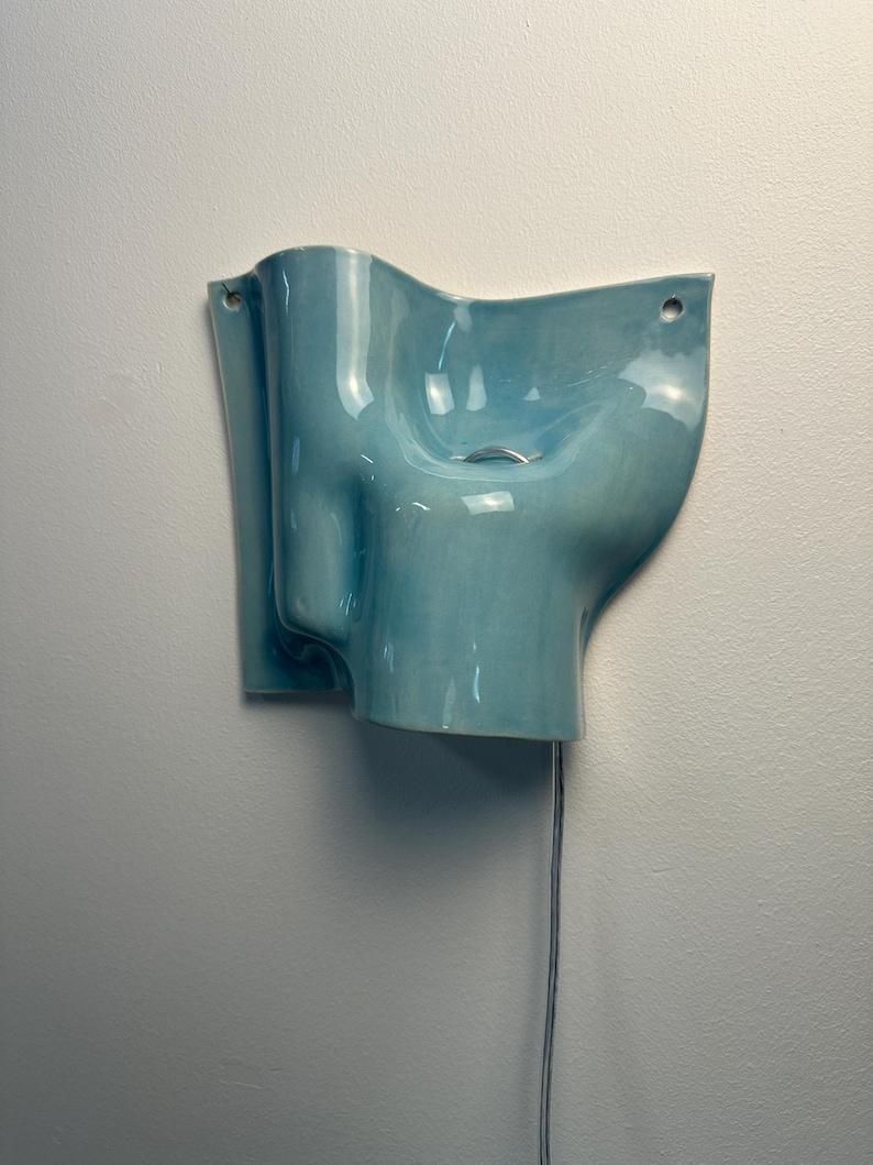 May include: A wall-mounted light blue ceramic wall light with a glossy finish and an organic, undulating shape. The fixture features two circular holes for mounting and a small silver-toned metal pull chain hanging from the centre for operation.