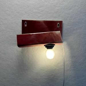 Może przedstawiać: Lampa ścienna z prostokątną, błyszczącą ciemnoczerwoną ceramiczną podstawą i zakrzywionym, wystającym elementem. Czarna oprawka mieści świecącą okrągłą żar&oacute;wkę, a z oprawy zwisa cienki biały przew&oacute;d zasilający.