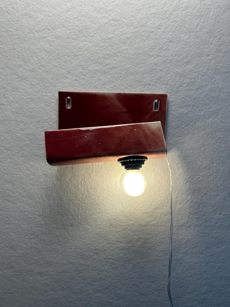 Może przedstawiać: Lampa ścienna z prostokątną, błyszczącą ciemnoczerwoną ceramiczną podstawą i zakrzywionym, wystającym elementem. Czarna oprawka mieści świecącą okrągłą żar&oacute;wkę, a z oprawy zwisa cienki biały przew&oacute;d zasilający.