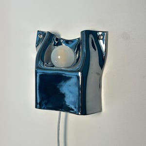 May include: A wall-mounted lamp featuring a reflective, chrome-finished, irregular ceramic housing with a smooth, undulating surface. A single, round, frosted white light bulb is centered within the recessed upper section. A thin, clear power cord extends downward.
