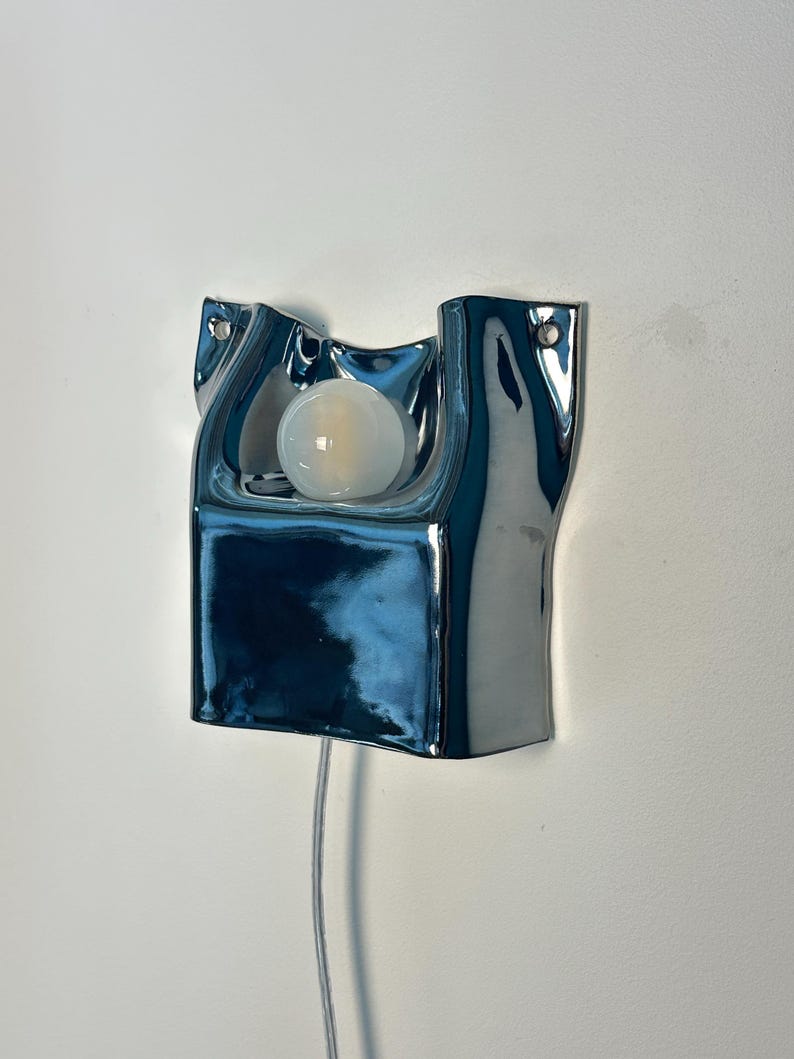 May include: A wall-mounted lamp featuring a reflective, chrome-finished, irregular ceramic housing with a smooth, undulating surface. A single, round, frosted white light bulb is centered within the recessed upper section. A thin, clear power cord extends downward.