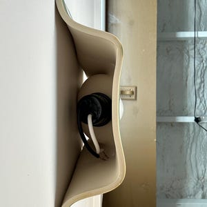 May include: A wall-mounted light fitting featuring a curved, beige ceramic shade with a wavy, organic silhouette. The fitting includes a black socket housing a white bulb, with black and white electrical wires extending from the centre.