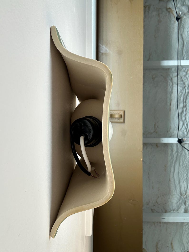 May include: A wall-mounted light fitting featuring a curved, beige ceramic shade with a wavy, organic silhouette. The fitting includes a black socket housing a white bulb, with black and white electrical wires extending from the centre.