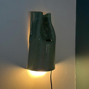 May include: A wall-mounted light fixture made of sage green ceramic with a speckled finish. The lamp has an irregular, organic shape with a hollow center and emits a warm, glowing light from the bottom, casting a soft illumination on the wall.