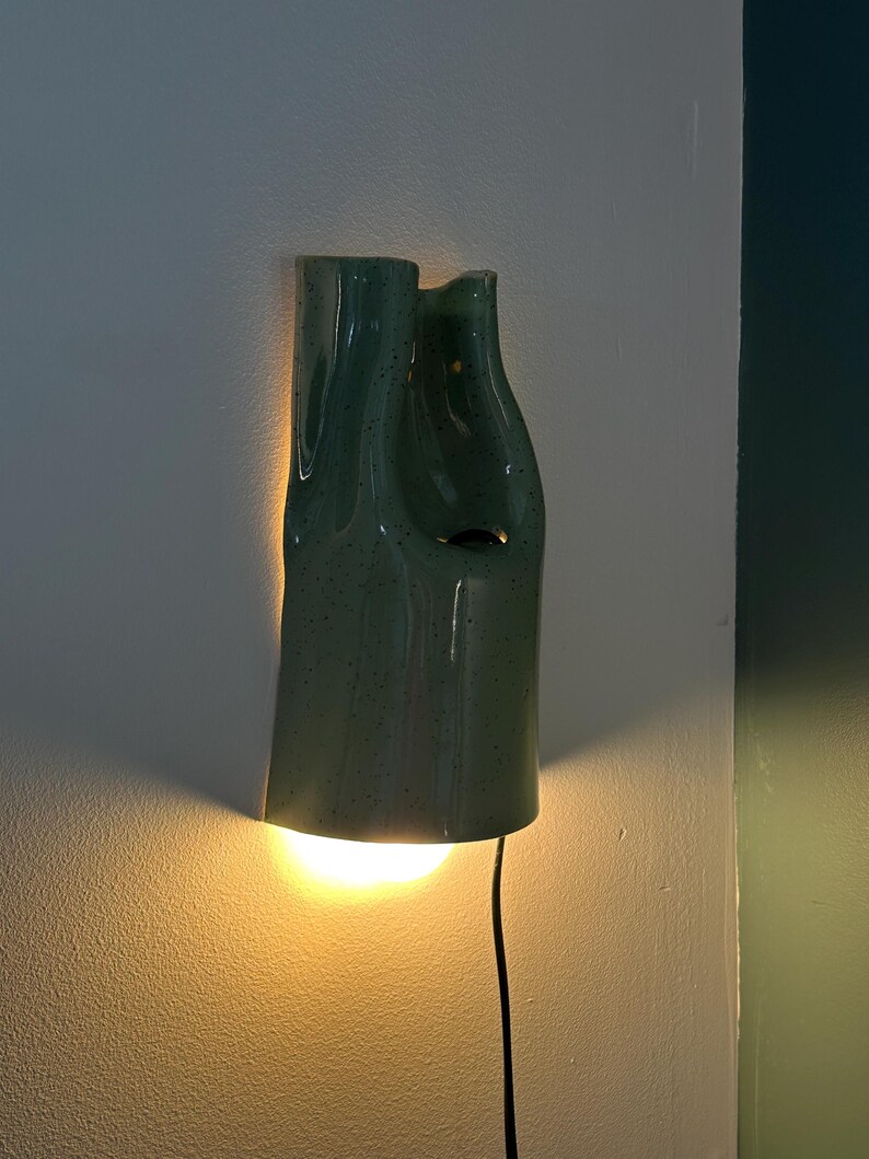 May include: A wall-mounted light fixture made of sage green ceramic with a speckled finish. The lamp has an irregular, organic shape with a hollow center and emits a warm, glowing light from the bottom, casting a soft illumination on the wall.