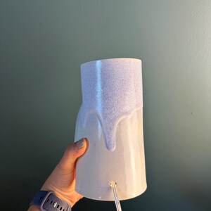 K&ouml;nnte beinhalten: Eine zylindrische Keramiklampe mit einem glatten, hellen Sockel und einer strukturierten, hellvioletten Glasur, die von oben herabl&auml;uft. Die Lampe hat eine kleine kreisf&ouml;rmige &Ouml;ffnung nahe der Unterseite, aus der ein transparentes Stromkabel ragt.