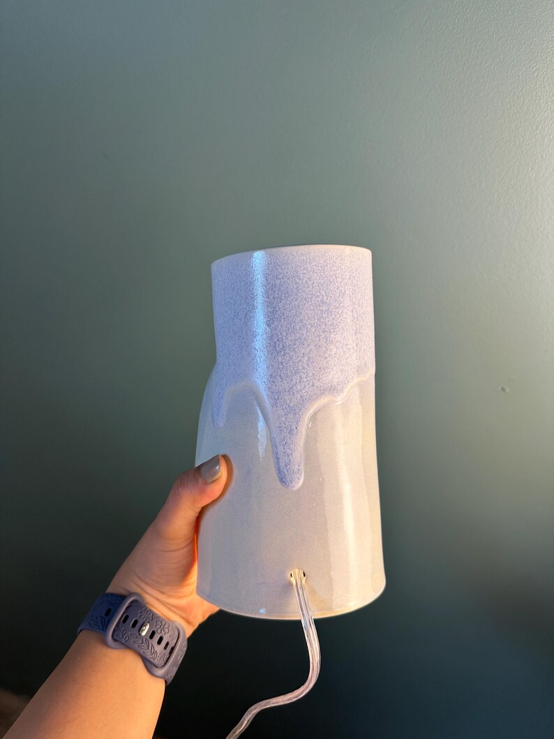 K&ouml;nnte beinhalten: Eine zylindrische Keramiklampe mit einem glatten, hellen Sockel und einer strukturierten, hellvioletten Glasur, die von oben herabl&auml;uft. Die Lampe hat eine kleine kreisf&ouml;rmige &Ouml;ffnung nahe der Unterseite, aus der ein transparentes Stromkabel ragt.
