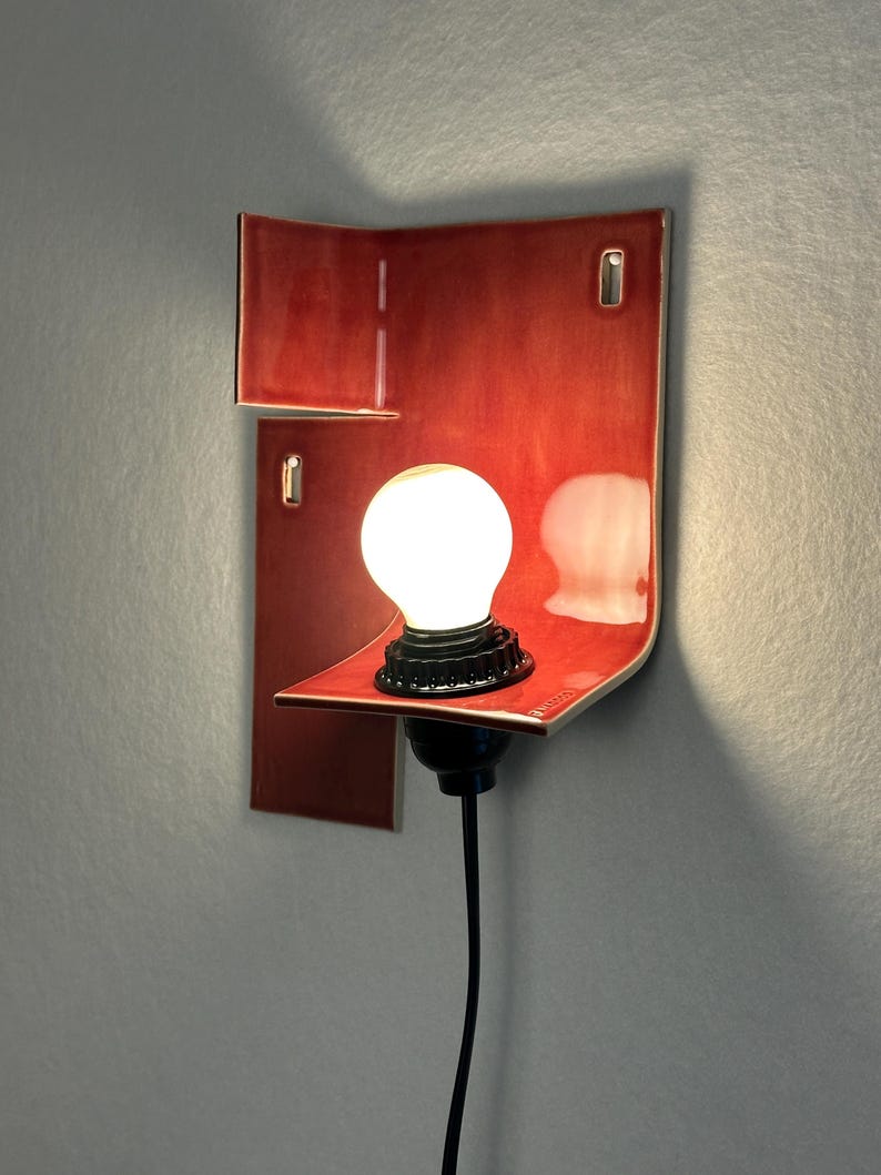 Pode incluir: Arandela de parede vermelha moderna com um design curvo. A arandela apresenta uma l&acirc;mpada redonda e brilhante e uma base preta. A lumin&aacute;ria &eacute; montada em uma parede cinza, mostrando uma est&eacute;tica minimalista.