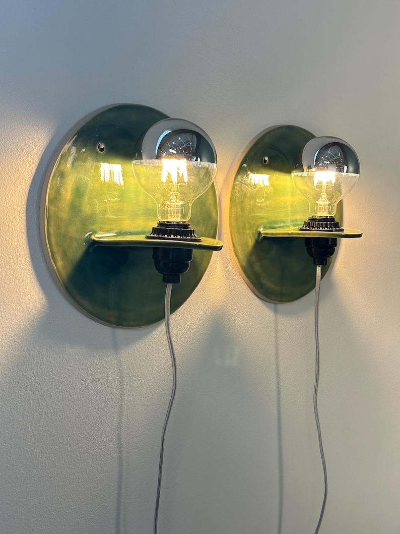 May include: Two wall-mounted sconces featuring circular, glossy green ceramic backplates with a small integrated shelf. Each fixture holds a clear glass light bulb with a silver-dipped top, a black socket, and a thin, dangling power cord.