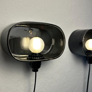 May include: Two black wall sconces with a glossy finish, each featuring a spherical light bulb. The sconces have a unique, curved design and are mounted on a light grey wall. The bulbs emit a warm, inviting glow.