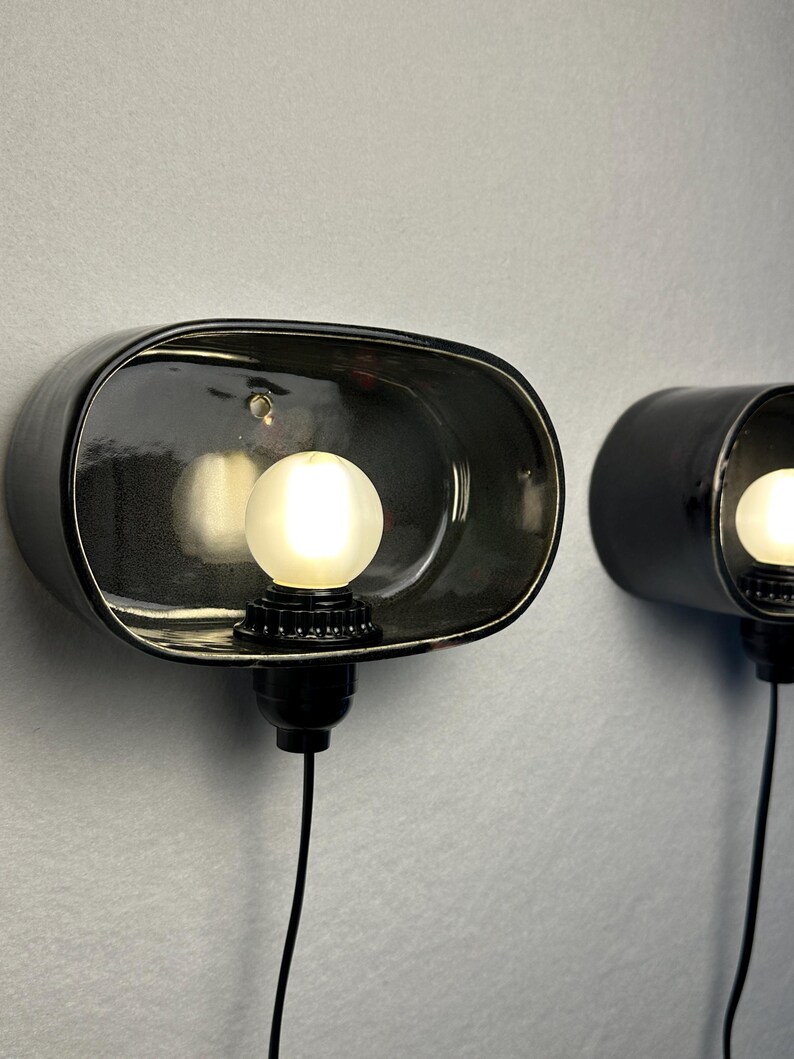 May include: Two black wall sconces with a glossy finish, each featuring a spherical light bulb. The sconces have a unique, curved design and are mounted on a light grey wall. The bulbs emit a warm, inviting glow.
