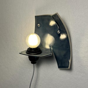 May include: A ceramic wall sconce in a dark blue glaze, featuring a shelf with a black light fixture and a glowing round bulb. The sconce has a unique, curved shape with visible mounting hardware.