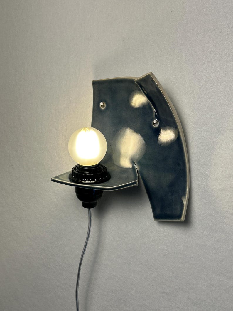 May include: A ceramic wall sconce in a dark blue glaze, featuring a shelf with a black light fixture and a glowing round bulb. The sconce has a unique, curved shape with visible mounting hardware.