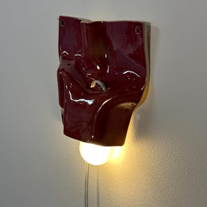 May include: A wall-mounted lamp featuring a glossy, deep red ceramic shade with an irregular, folded form. The fixture includes a circular light bulb emitting a warm glow from the bottom, a small metal hook in the center, and a thin power cord.
