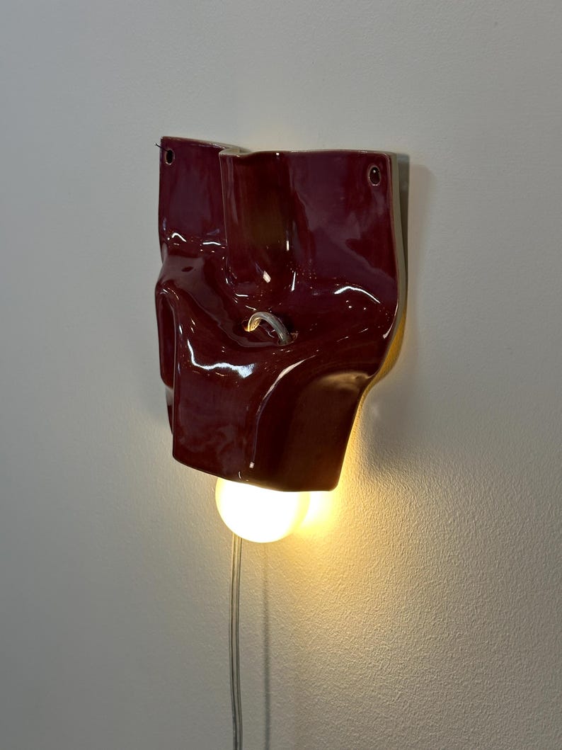 May include: A wall-mounted lamp featuring a glossy, deep red ceramic shade with an irregular, folded form. The fixture includes a circular light bulb emitting a warm glow from the bottom, a small metal hook in the center, and a thin power cord.