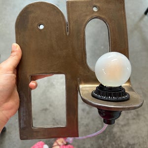 K&ouml;nnte beinhalten: Eine Wandleuchte mit einer bronzefarbenen Metallplatte, die einen bogenf&ouml;rmigen und einen rechteckigen Ausschnitt aufweist. Eine schwarze Fassung h&auml;lt eine runde, mattwei&szlig;e Gl&uuml;hbirne, und ein d&uuml;nnes wei&szlig;es Stromkabel f&uuml;hrt aus der Basis.