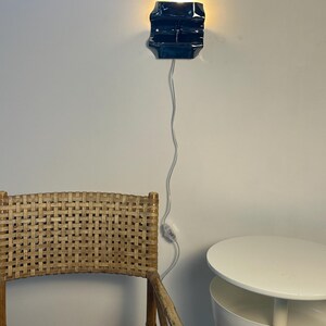 May include: A dark blue, rectangular wall-mounted lamp with a glossy finish, emitting a warm light from the top and bottom. The lamp is connected to a thin white power cord that hangs vertically down the wall, featuring an inline toggle switch.