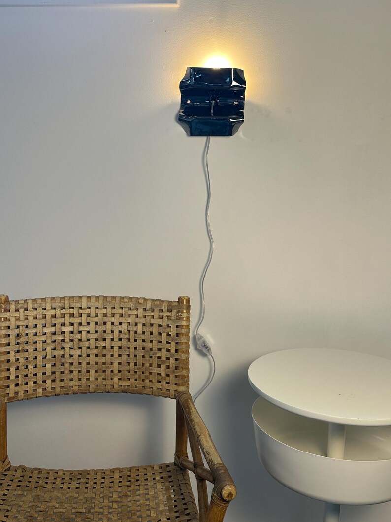 May include: A dark blue, rectangular wall-mounted lamp with a glossy finish, emitting a warm light from the top and bottom. The lamp is connected to a thin white power cord that hangs vertically down the wall, featuring an inline toggle switch.