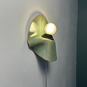 Puede incluir: Una l&aacute;mpara de pared con una pantalla de cer&aacute;mica verde claro curvada con motas oscuras. El aplique incluye un casquillo negro que sostiene una bombilla blanca esf&eacute;rica e iluminada. Un cable de alimentaci&oacute;n fino cuelga de la base.