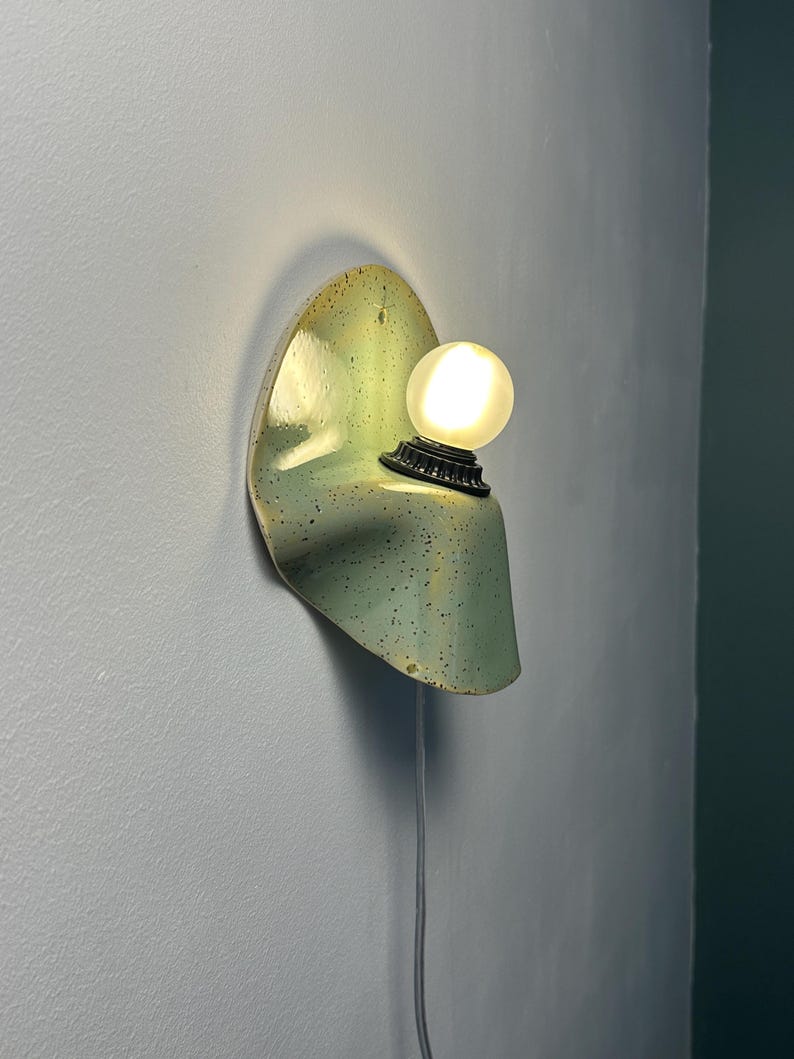 Puede incluir: Una l&aacute;mpara de pared con una pantalla de cer&aacute;mica verde claro curvada con motas oscuras. El aplique incluye un casquillo negro que sostiene una bombilla blanca esf&eacute;rica e iluminada. Un cable de alimentaci&oacute;n fino cuelga de la base.