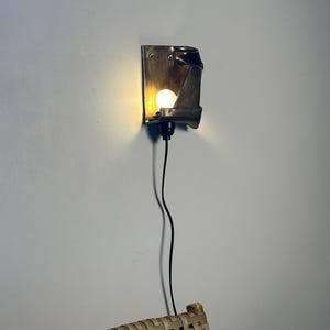 May include: A wall-mounted sconce featuring a dark metallic, folded rectangular backplate with a single glowing bulb. The fixture is connected to a thin black power cord that hangs vertically downward from the base of the light socket.