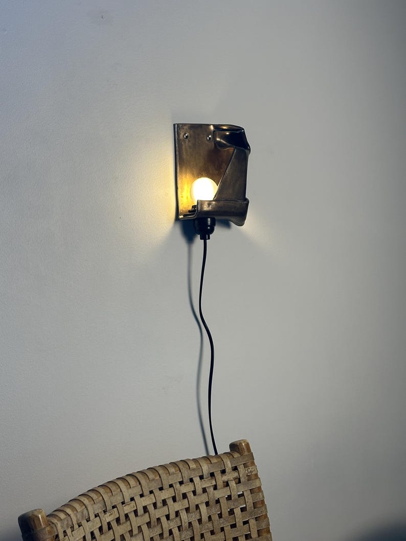 May include: A wall-mounted sconce featuring a dark metallic, folded rectangular backplate with a single glowing bulb. The fixture is connected to a thin black power cord that hangs vertically downward from the base of the light socket.
