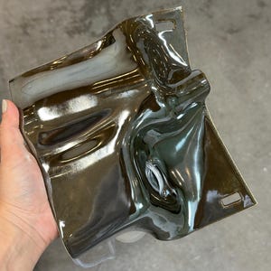 May include: A rectangular ceramic wall plate with a high-gloss, dark metallic brown glaze. The surface features an undulating, fluid texture with deep curves and indentations. Two small rectangular mounting slots are positioned at the top and bottom.