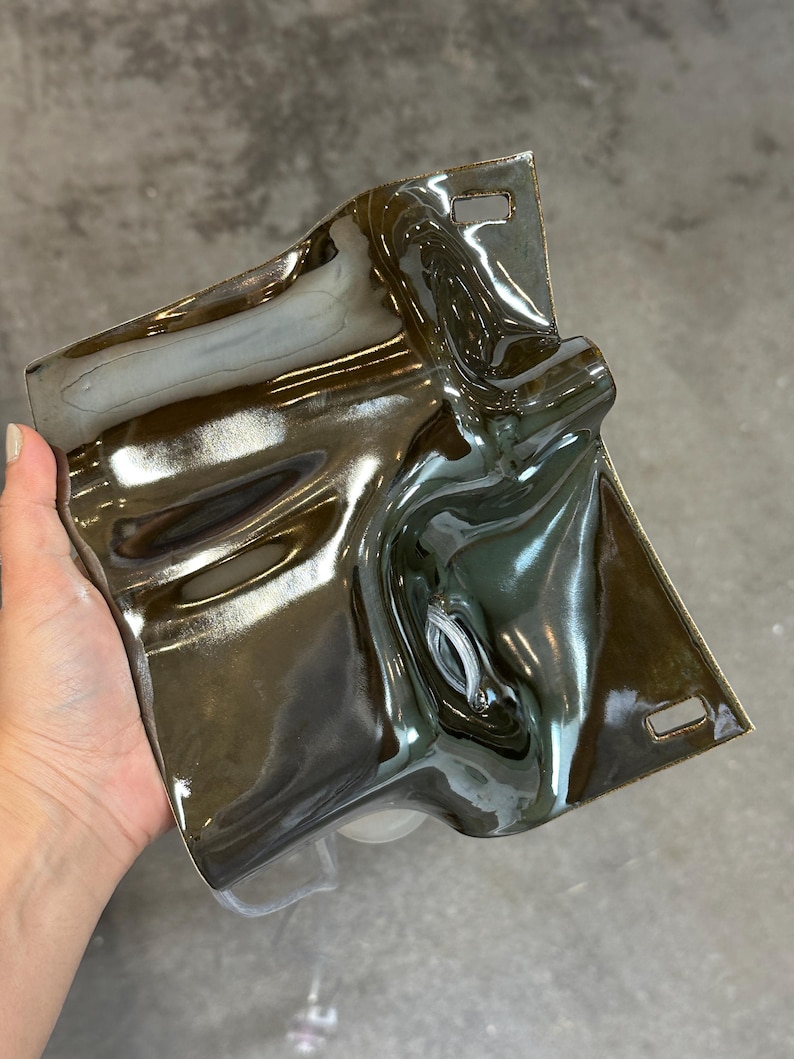 May include: A rectangular ceramic wall plate with a high-gloss, dark metallic brown glaze. The surface features an undulating, fluid texture with deep curves and indentations. Two small rectangular mounting slots are positioned at the top and bottom.
