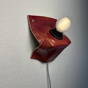 Puede incluir: Un aplique de pared con una pantalla de cer&aacute;mica de color rojo oscuro brillante y forma escultural plegada. La l&aacute;mpara incluye un casquillo negro con una bombilla redonda de cristal esmerilado blanco y un cable fino en la parte inferior.