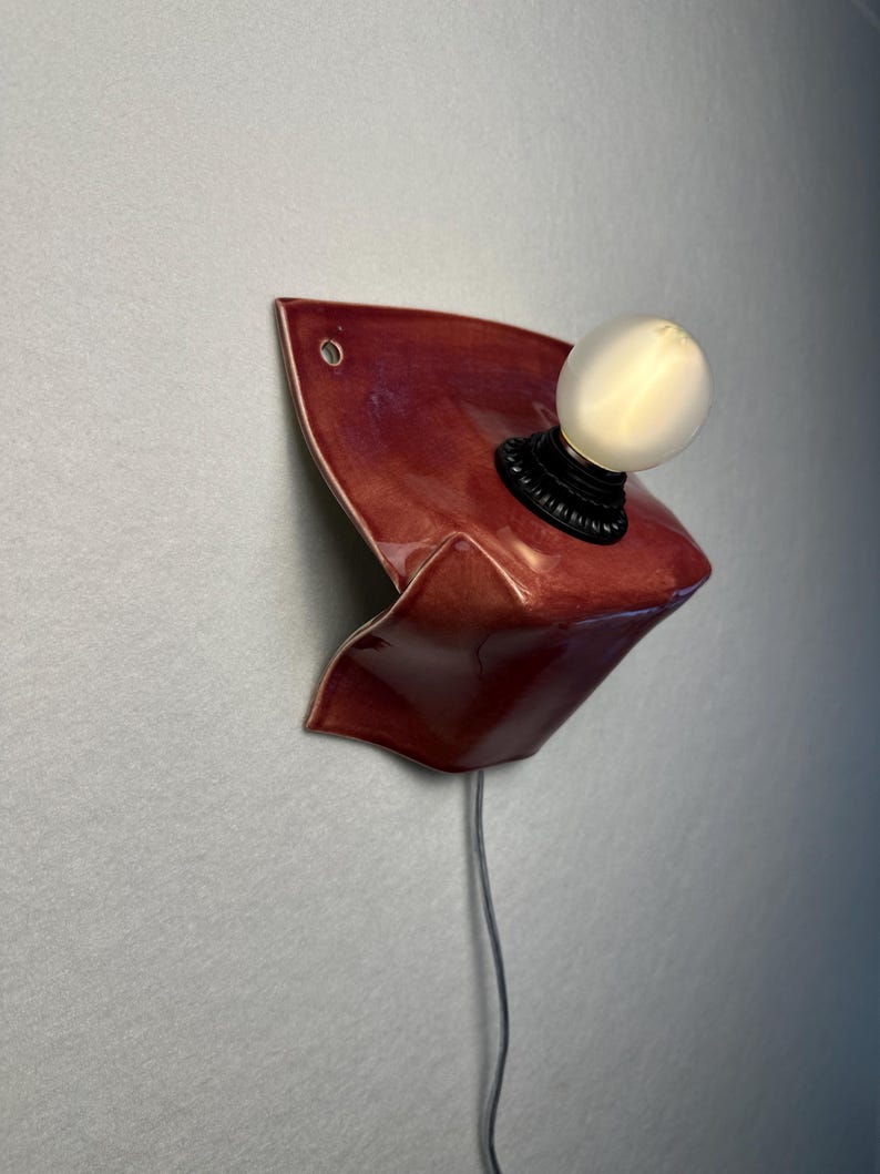 Puede incluir: Un aplique de pared con una pantalla de cer&aacute;mica de color rojo oscuro brillante y forma escultural plegada. La l&aacute;mpara incluye un casquillo negro con una bombilla redonda de cristal esmerilado blanco y un cable fino en la parte inferior.